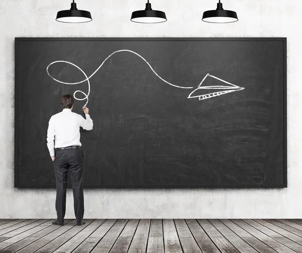 Rear view of a man who is drawing an arrow as a concept of possible solution of the complicated problem. Black chalkboard, wooden floor and three black veiling lights are in the room. — Stock Photo, Image