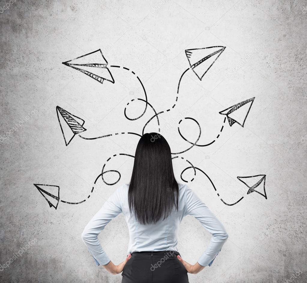 Rear view of brunette in formal clothes who is pondering about possible solutions of the complicated problem. Many arrows with different directions are drawn around her head on the concrete wall.