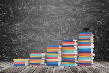 A stair is made of colourful books. A graduation hat is on the final step. Black chalk board with math formulas on the background. Wooden floor.