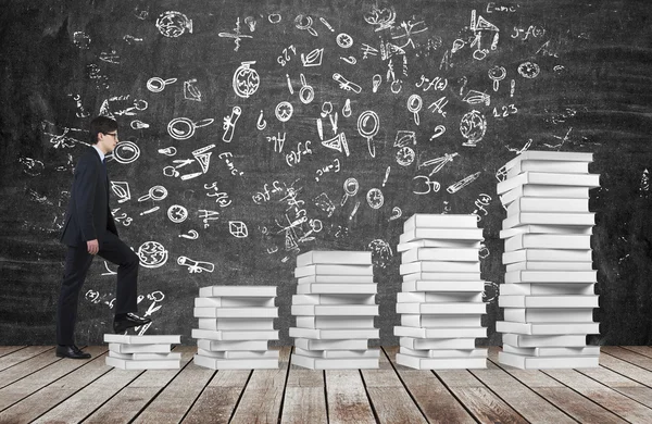Ein Mann geht eine Treppe hinauf, die aus weißen Büchern besteht. Bildungsikonen sind auf die schwarze Tafel gemalt. Holzboden. — Stockfoto