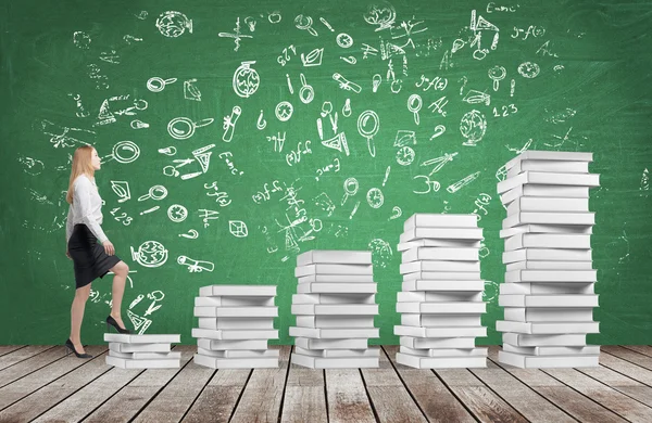 A woman is going up using a stairs which are made of white books. Educational icons are drawn on the green chalkboard. Wooden floor. — Stock Photo, Image