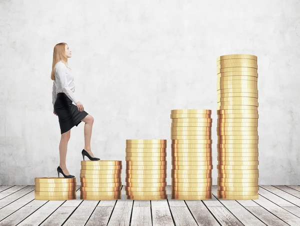 A woman in formal clothes is going up using a stairs which are made of golden coins. A concept of success. Concrete wall and wooden floor. — Stock Photo, Image