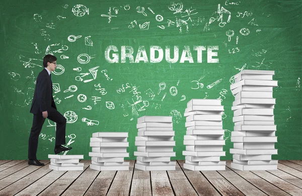 A man is going up using a stairs which are made of white books. The word graduate is on the green chalkboard. Wooden floor. — Stockfoto