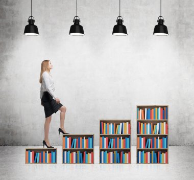 A woman in formal clothes is going up on the bookshelf. A concept of different level of education. Concrete room with four black ceiling lights.
