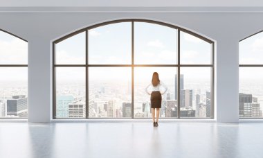 Rear view of a woman in formal clothes who is looking out the window in a modern clean interior with huge panoramic windows. New York city view. Toned Image.