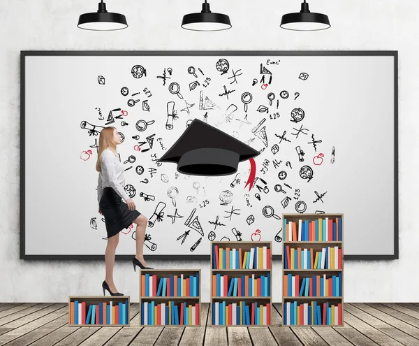 A woman in formal clothes is going up on the bookshelf. A concept of different level of education. A sketched graduation hat and different educational icons on the whiteboard. — Stockfoto