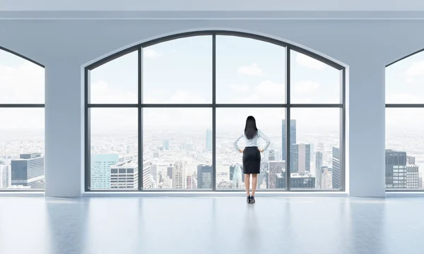 Rear view of a woman in formal clothes who is looking out the window in a modern clean interior with huge panoramic windows. New York city view. — Stock Photo, Image