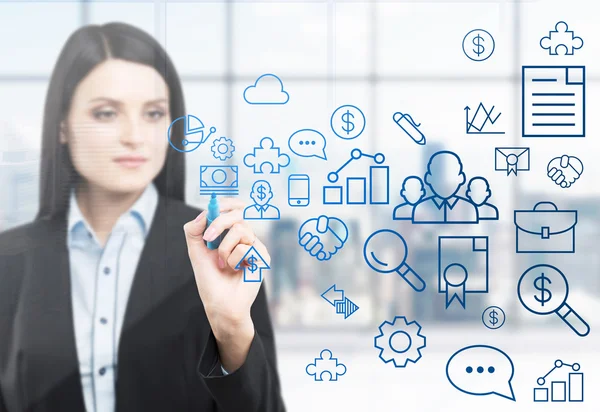 A brunette woman is drawing some business icons on the glass screen. Modern panoramic office with New York view in blur on the background. — Stok fotoğraf