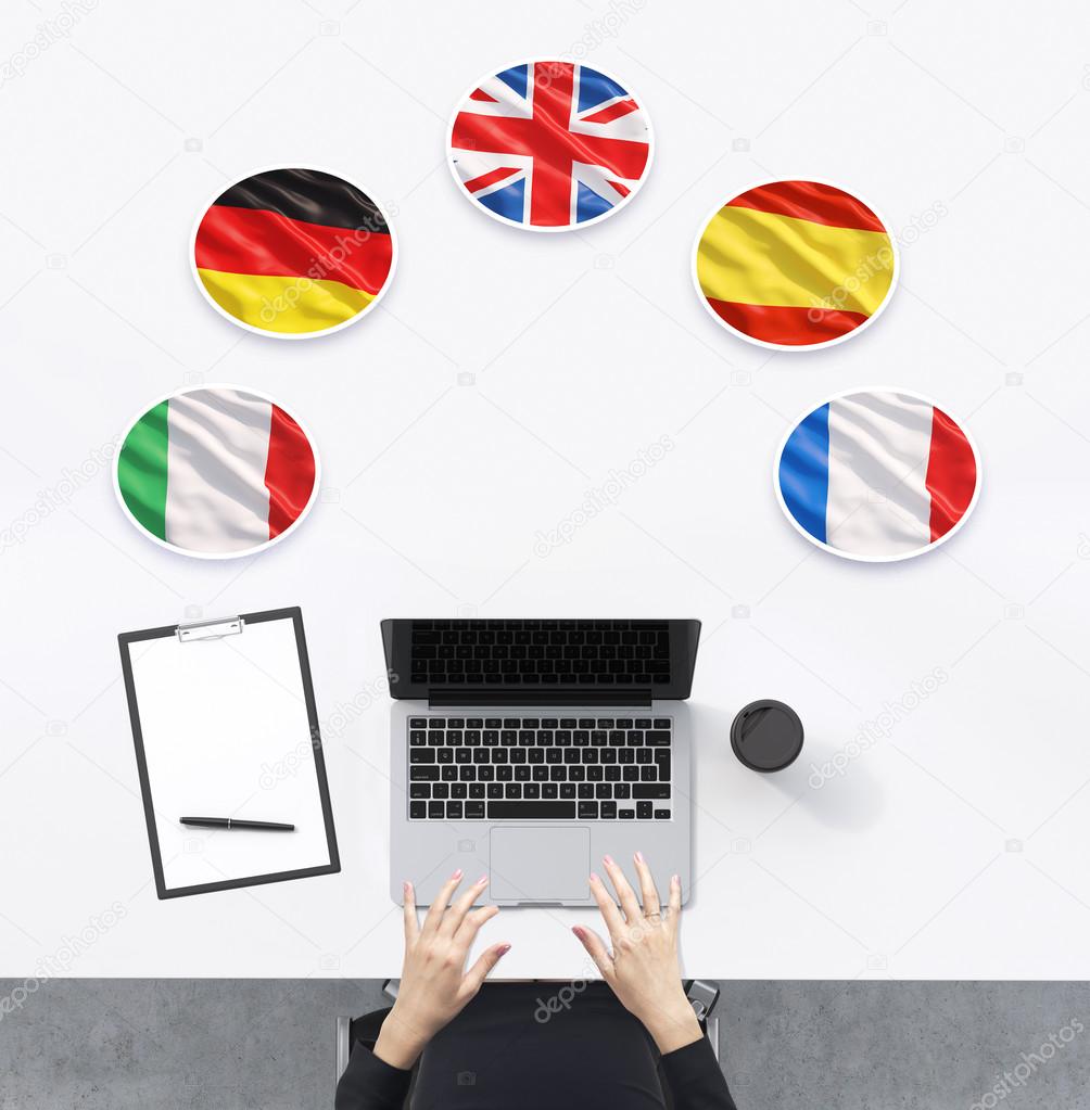 Top view of the working place with woman's hands. A laptop, notepad and a cup of coffee are on the table. Italian, German, United Kingdom, Spanish and French flags in the bubbles.