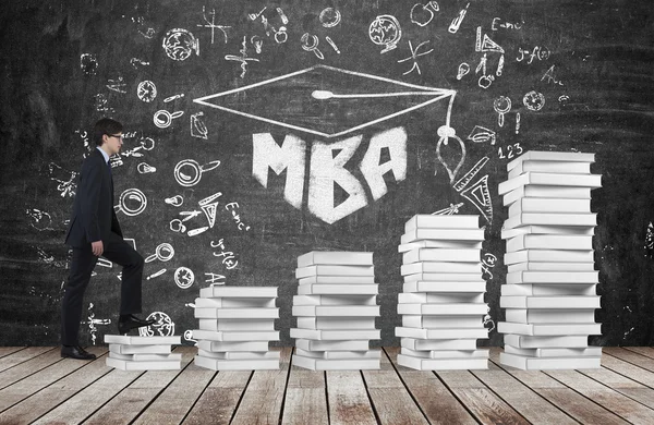 Un hombre está subiendo usando unas escaleras que están hechas de libros blancos para alcanzar el sombrero de graduación. La palabra escrita MBA se dibuja en la pizarra negra que simboliza una educación profesional de negocios . — Foto de Stock