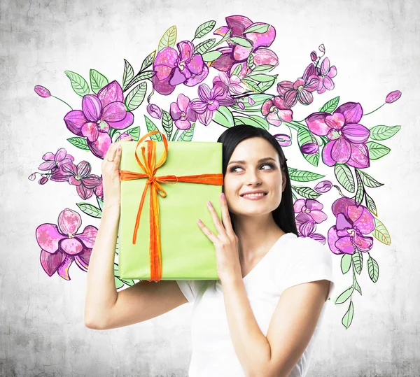 Versucht eine neugierige brünette Frau zu erraten, was sich in der grünen Geschenkbox befindet. die Skizze von lila Blumen wird auf die Betonwand gezeichnet. — Stockfoto