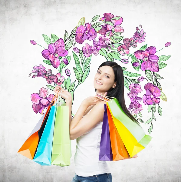 Kleurrijke tassen houdt een gelukkig brunette vrouw van mooie winkels. Het concept van het winkelen. Een schets van paarse bloemen wordt op de betonnen muur getekend. — Stockfoto