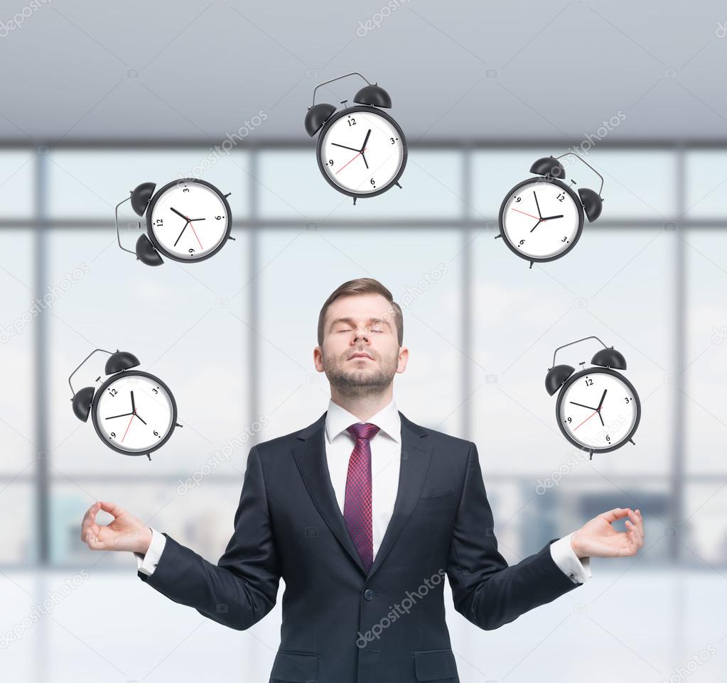 Meditative businessman is pondering about time management in the modern panoramic office. The person in formal suit is surrounded by alarm clocks. A concept of time management and deadlines.