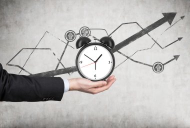 A businessman's hand holds an alarm clock. There is a growing line charts behind the alarm clock. A concept of time management or billing services in legal or consulting company.