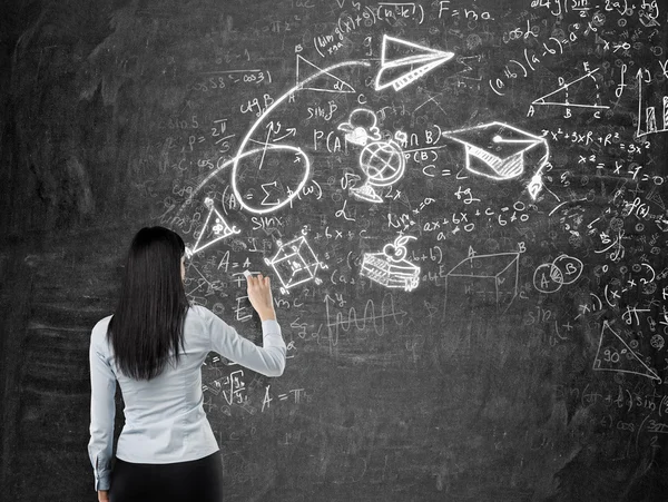 Visão traseira de uma senhora morena que está desenhando um esboço de formatura. Fórmulas matemáticas, uma seta e figuras geométricas são desenhadas no quadro negro . — Fotografia de Stock