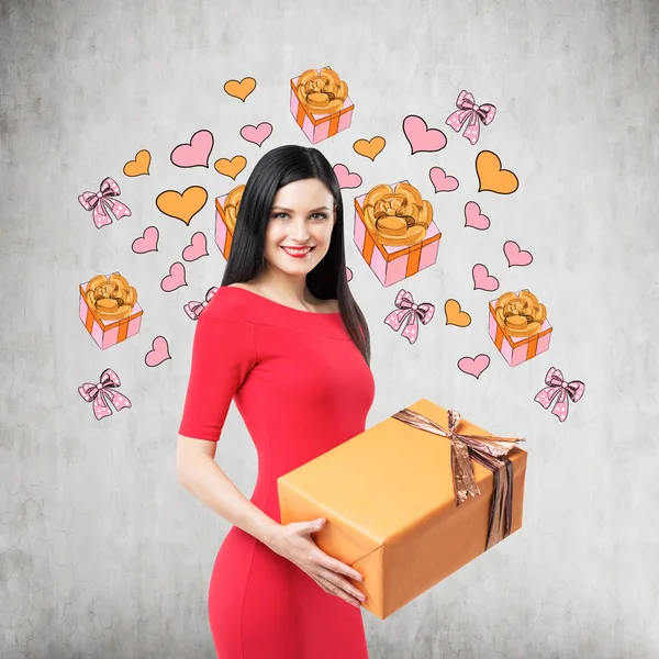 A brunette in red dress holds an orange gift box. Gift and heart icons are drawn on the concrete wall. — Stockfoto