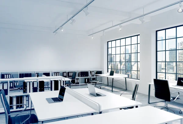 Workplaces in a bright modern loft open space office. Tables equipped with laptops; corporate documents' shelves. New York in the panoramic windows. 3D rendering.