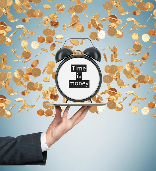 A hand holds a tablet with alarm clock. Golden coins are falling down from the ceiling. The concept of time is money. Light blue background. — Stockfoto