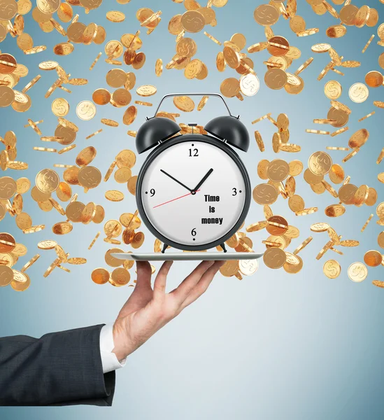 A hand holds a tablet with alarm clock. Golden coins are falling down from the ceiling. The concept of time is money. Light blue background. — Stock Photo, Image