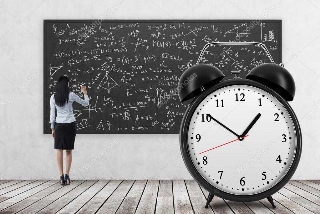 Rear view of business lady who is writing math formulas on the black chalkboard. The huge alarm clock is on the foreground. A concept of time and quantitative science. Wooden floor and concrete wall.