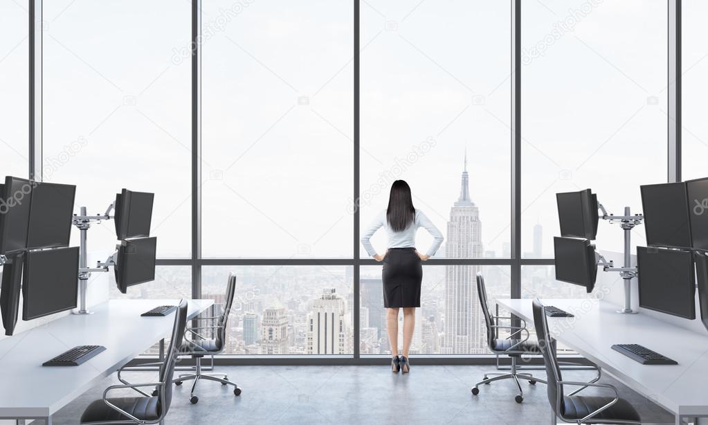 Rear view of a lady in formal suit who is looking out the window in the modern panoramic office with New York view. White tables equipped with modern trader's stations and black chairs.