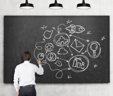 Rear view of young man in formal clothes who is drawing connected business icons on the black chalkboard. The concept of a communication building a relationships.