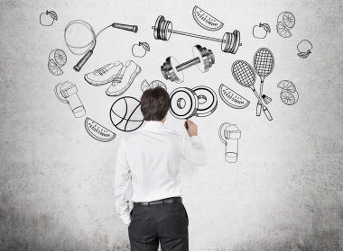 A rear view of a person in formal clothes who is drawing a range of sport activities and sport equipment on the concrete wall. A concept of gym culture among office clerks.