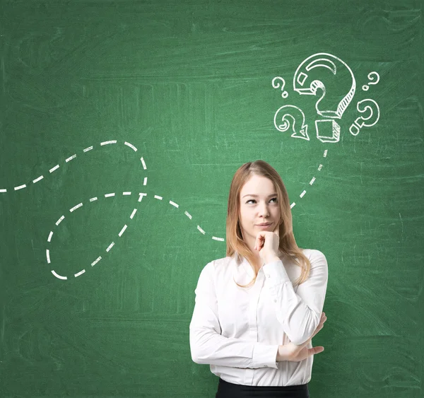 A young beautiful lady in formal clothes is thinking about unanswered questions. Question marks are drawn on the green chalkboard.