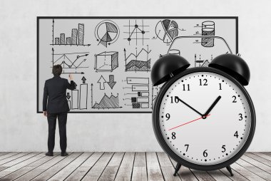 Rear view of businessman who is drawing analytical charts on the whiteboard. The huge alarm clock is on the foreground. A concept of time and analyst researches. Wooden floor and concrete wall.