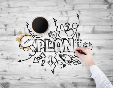 A top view of a cup of coffee and a hand which is drawing a sketch of developing a new business plan. A wooden surface as a background.