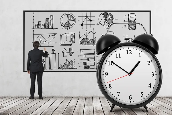 Rear view of businessman who is drawing analytical charts on the whiteboard. The huge alarm clock is on the foreground. A concept of time and analyst researches. Wooden floor and concrete wall. — Stock Photo, Image