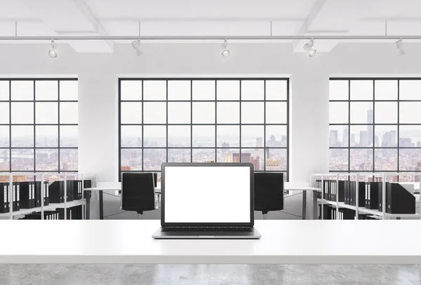 A workplace in a bright modern loft open space office. A working desk is equipped with a modern laptop with white copy space in the screen. Docs shelves. New York view in the windows. 3D rendering. — стокове фото