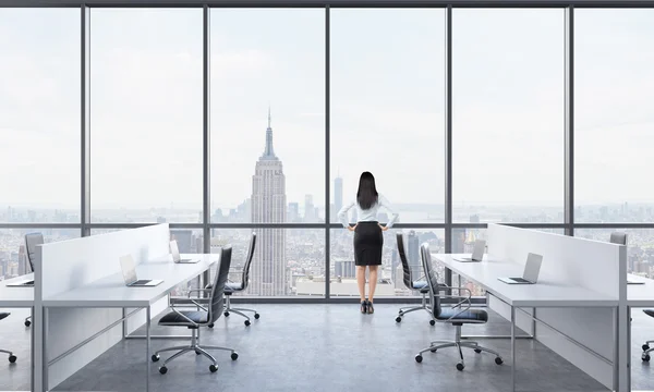 Rear view of a brunette who is looking out the window in the modern panoramic office with New York view. White tables equipped with modern laptops and black chairs. — 스톡 사진