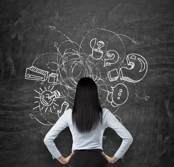 Rear view of brunette who is looking at the black chalkboard with drawn sketch of business icons, exclamation and question marks. The concept of brainstorm process. — Stockfoto