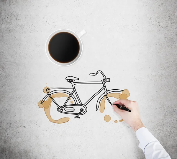 Top view of a cup of coffee and a drawing process of a bicycle on the concrete surface. A hand in formal white shirt with a pen. — Stockfoto