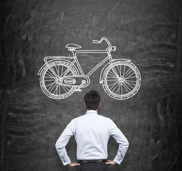 Visão traseira de um homem de negócios em roupas formais que está olhando para o enorme quadro negro com um esboço desenhado de uma bicicleta. Um conceito de modos ecológicos de deslocação e de viagem . — Fotografia de Stock