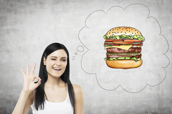 A brunette woman is thinking about burger. A fast food concept. Concrete background. — Stock Fotó