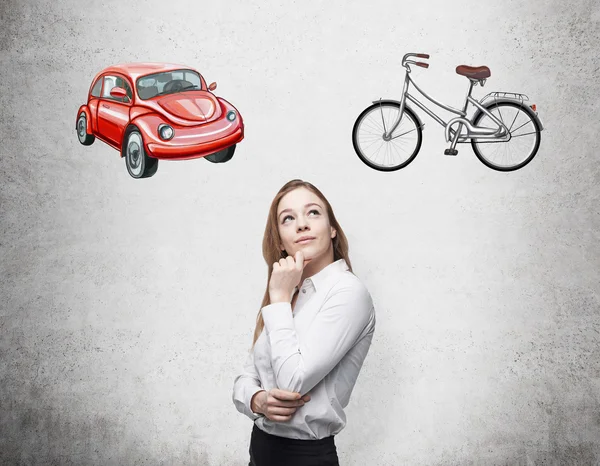 A beautiful woman is trying to chose the most suitable way for travelling or commuting. Two sketches of a car and a bicycle are drawn on the concrete wall. — Stock Photo, Image