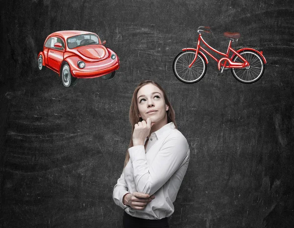 Uma mulher bonita está tentando escolher a maneira mais adequada para viajar ou viajar. Dois esboços de um carro e uma bicicleta são desenhados no fundo do quadro preto . — Fotografia de Stock