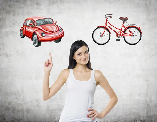 A beautiful woman is trying to chose the most suitable way for travelling or commuting. Her choice is a car. Two sketches of a car and a bicycle are drawn on the concrete wall. — Stock Photo, Image