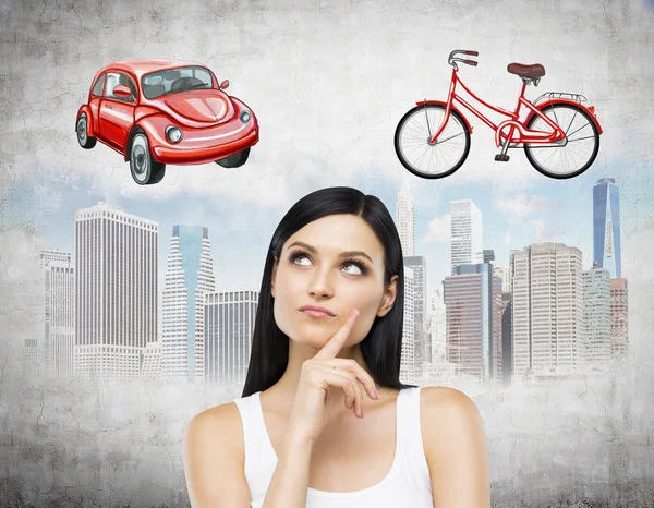 A brunette lady is trying to chose the most suitable way for travelling or commuting in the city. Sketches of a car, a bicycle and New York city are on the concrete background. — Stock Photo, Image