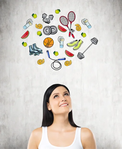 Pondering beautiful lady is thinking about her choice of sport activity. Colourful sport icons are drawn on the concrete wall. A concept of a healthy lifestyle. — Stock Photo, Image