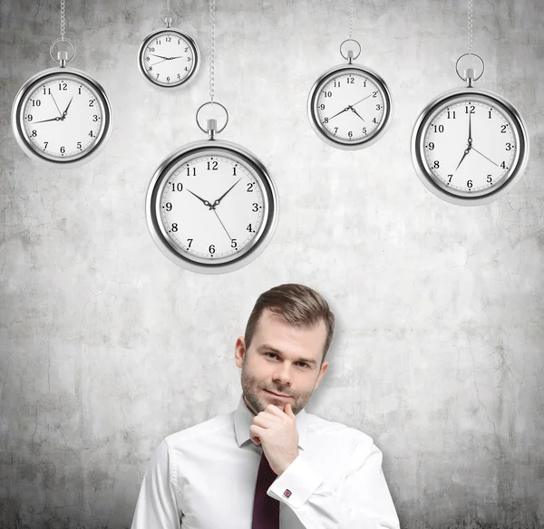 A portrait of a thinking handsome businessman who is holding his own chin. Several models of pocket watches are hovering in the air. A concept of a value of time in business. Concrete background. — Zdjęcie stockowe