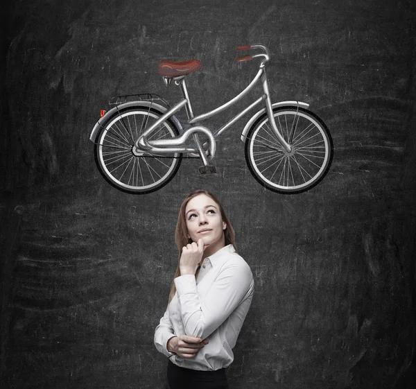 Una hermosa chica en ropa formal está soñando con una bicicleta nueva. Un boceto de una bicicleta se dibuja en la pizarra negra . —  Fotos de Stock