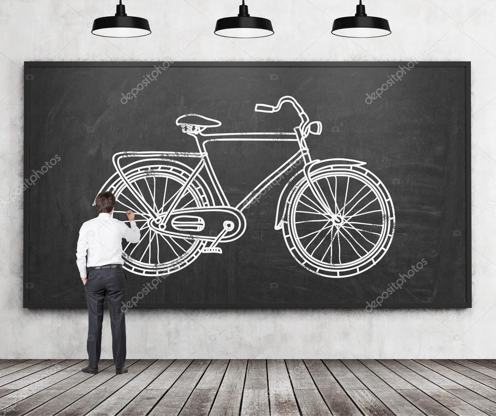 Rear view of a businessman in formal clothes who is drawing a sketch of a bicycle on the huge black chalkboard. A concept of environmental friendly ways of commuting or travelling.