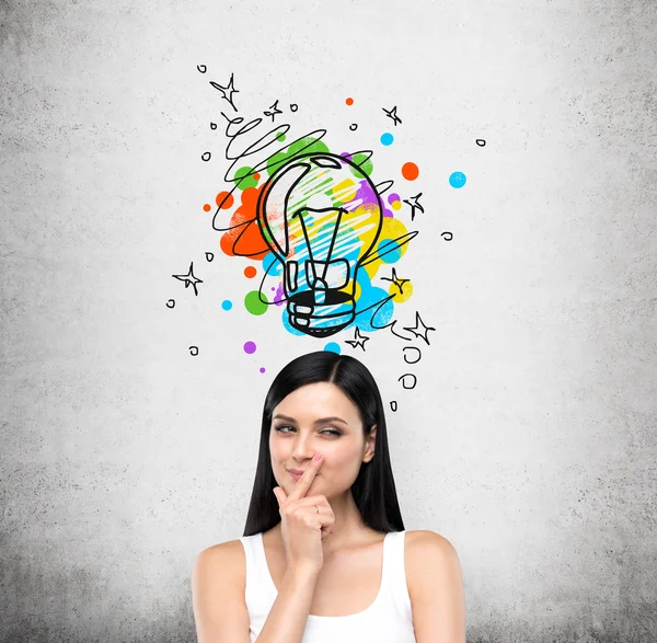 A portrait of a young artful brunette lady who is trying to create a new idea for some business project or case study. A colourful lightbulb as a concept of a new idea is drawn on the concrete wall. — Stock Photo, Image