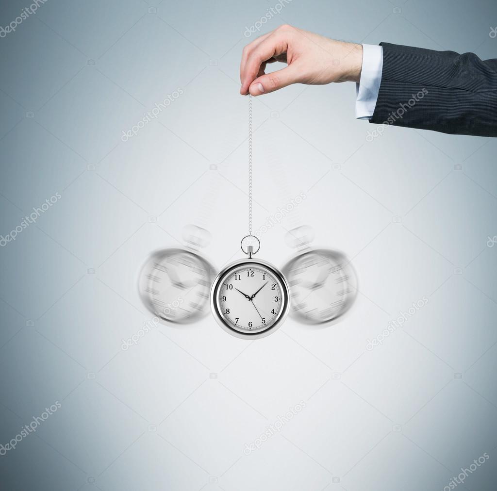 A hand holds a pocket watch in a chain as a pendulum. Light blue background. Time is money concept.