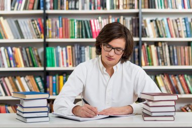 serious young man with dark hair sitting at a desk in the librar clipart