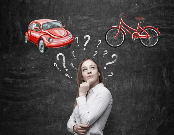 A beautiful woman is trying to chose the most suitable way for travelling or commuting. Two sketches of a car and a bicycle are drawn on the black chalkboard. — Stock Photo, Image