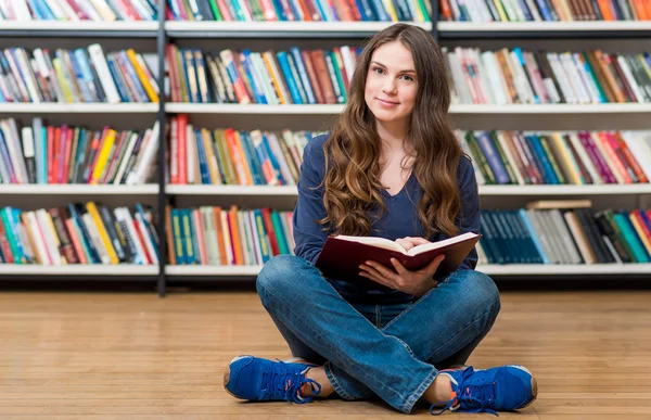 Sonriente joven sentada en el suelo en la biblioteca con cros —  Fotos de Stock